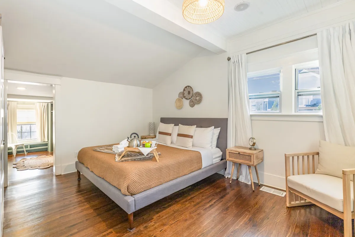 A serene bedroom with a bed dressed in decorative pillows, a wooden side table, a woven light fixture, and a window streaming in gentle sunlight. The overall vibe is tranquil and comfortable, making it an ideal space to recharge.