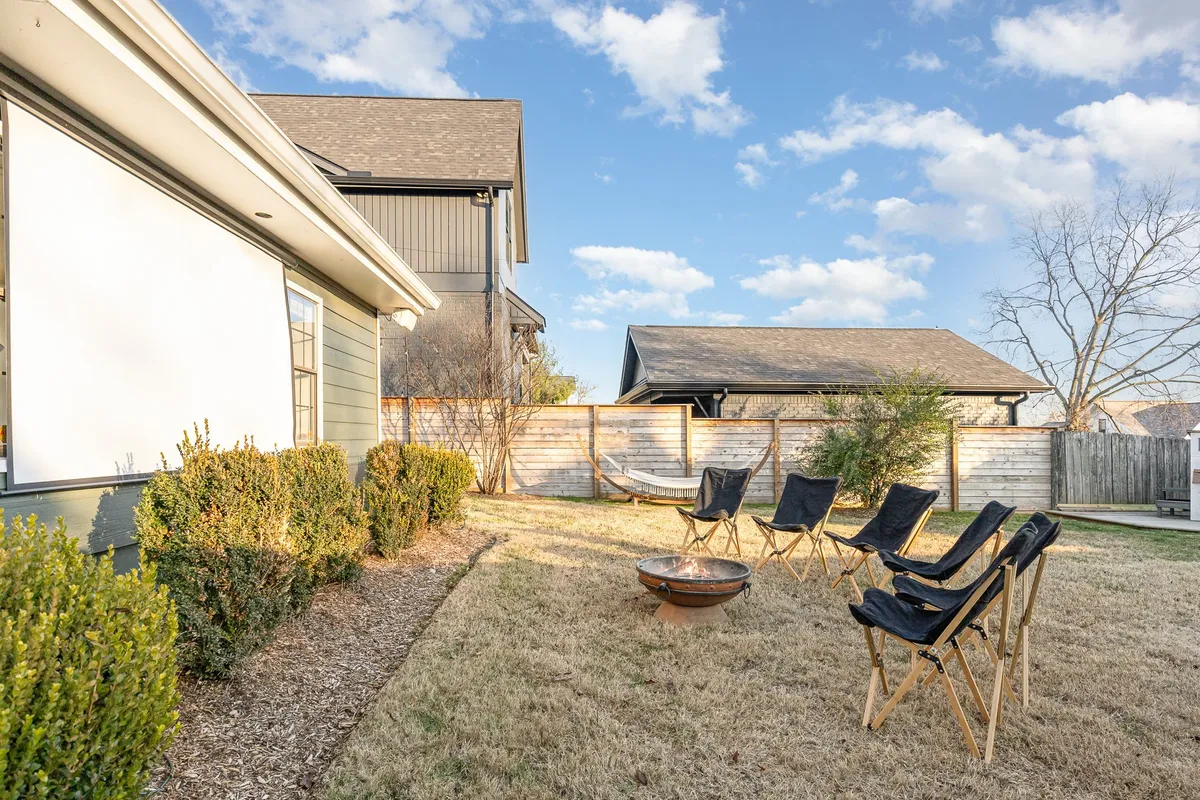 A lush backyard scene with a fire pit surrounded by lounge chairs, a hammock strung between trees, and landscaped bushes. The space is designed for socializing, relaxing, and outdoor conversations, ideal for both adults and children.