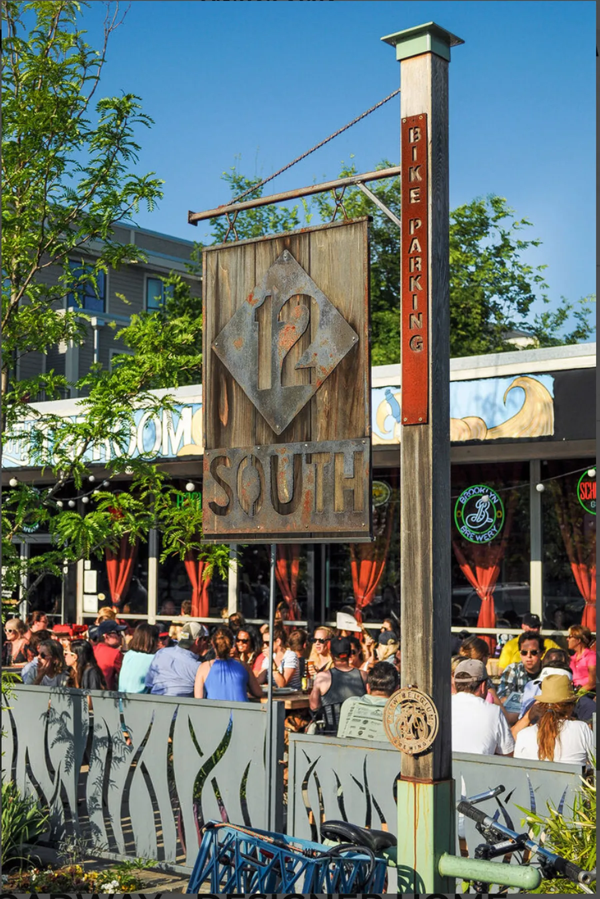 A vibrant scene of the 12 South neighborhood featuring a prominent '12 South' sign, crowds of people dining and socializing outdoors, ample seating, and a bike parking sign. The atmosphere is energetic and community-focused, conveying the area's popularity for food, drink, and social connection.