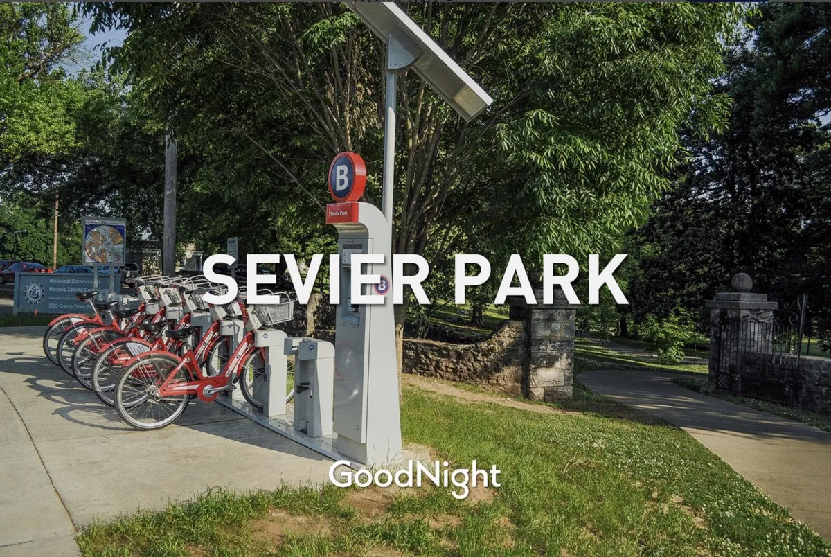 This image features the Sevier Park sign, a bicycle rental station, and the surrounding greenery, capturing a sunny and energetic outdoor atmosphere. It depicts a popular family-friendly space where children can play, families can walk, and locals can picnic.