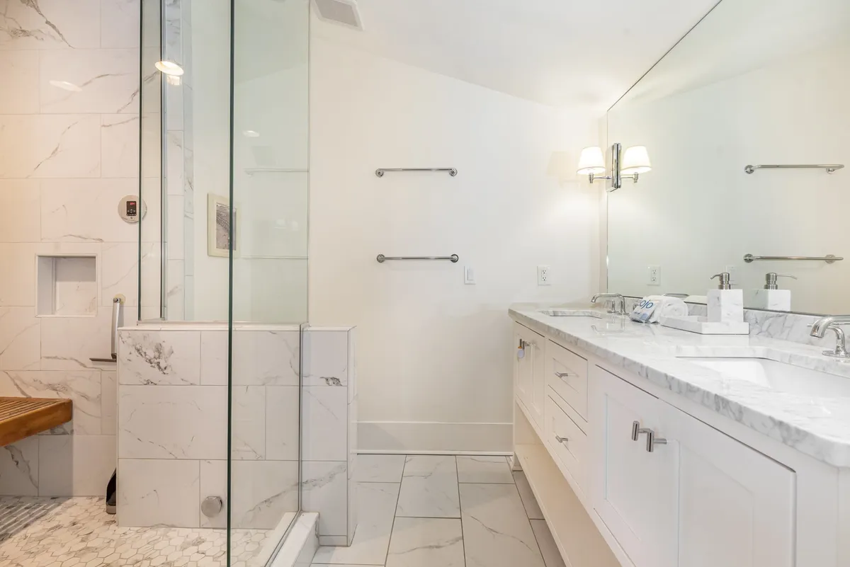 A modern bathroom featuring a glass shower enclosure, double sink vanity with a marble countertop, a wooden bench, and a large mirror. The space feels fresh, spacious, and well-equipped for both quick routines and relaxing baths.