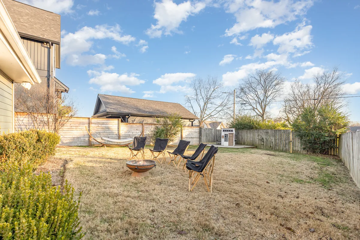 A cozy backyard scene featuring a hammock strung between trees, several lounge chairs encircling a fire pit, and a backdrop of leafy trees and a wooden fence. The setting is informal and welcoming, ideal for relaxation and family gatherings.