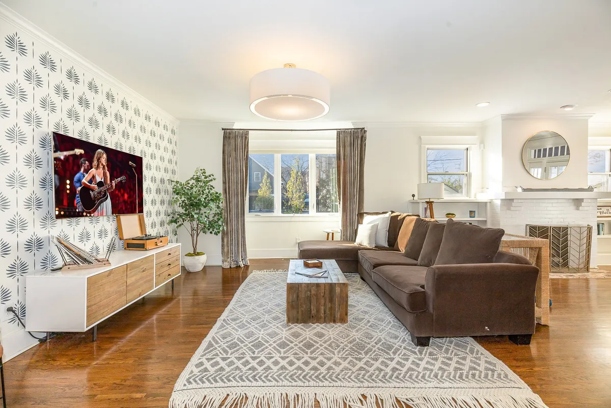 The image features a spacious, sunlit living room with a large sectional sofa arranged around a modern coffee table. Decorative plants add warmth and life to the space, while a large TV and entertainment center invite guests to relax, watch movies, or gather for memorable evenings. The atmosphere is both cozy and stylish, perfect for family groups or friends to unwind and connect.
