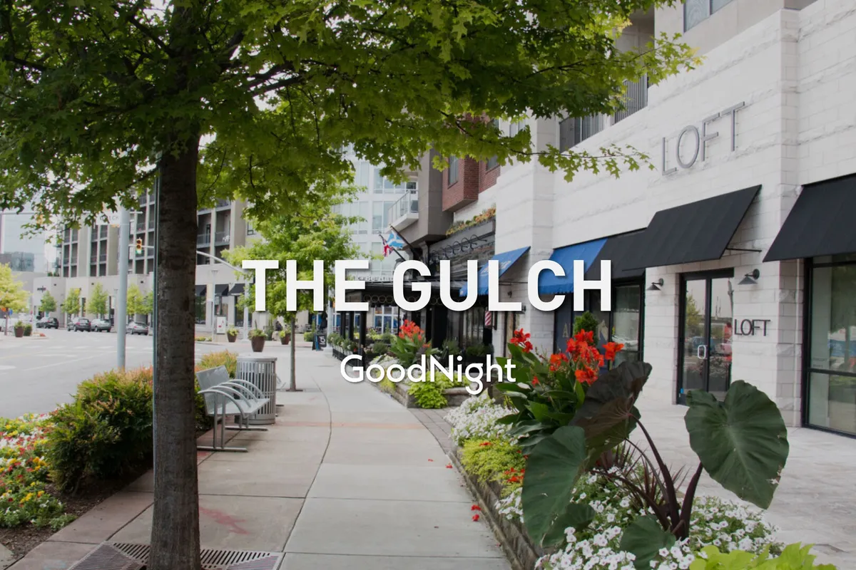 A vibrant, tree-lined sidewalk bordered by colorful flower beds, inviting storefronts, and benches. The scene is lively, with people strolling, shopping, and socializing, reflecting the neighborhood's walkability and pet-friendly character.
