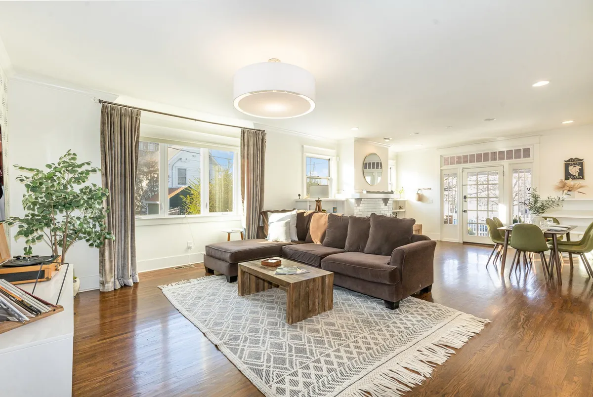 A bright and inviting open-plan living area featuring a large sectional sofa centered around a coffee table, with abundant natural light streaming through windows. In the background, the dining area is visible, seamlessly connecting the living and dining spaces. The space exudes comfort and warmth, ideal for group gatherings and shared moments.